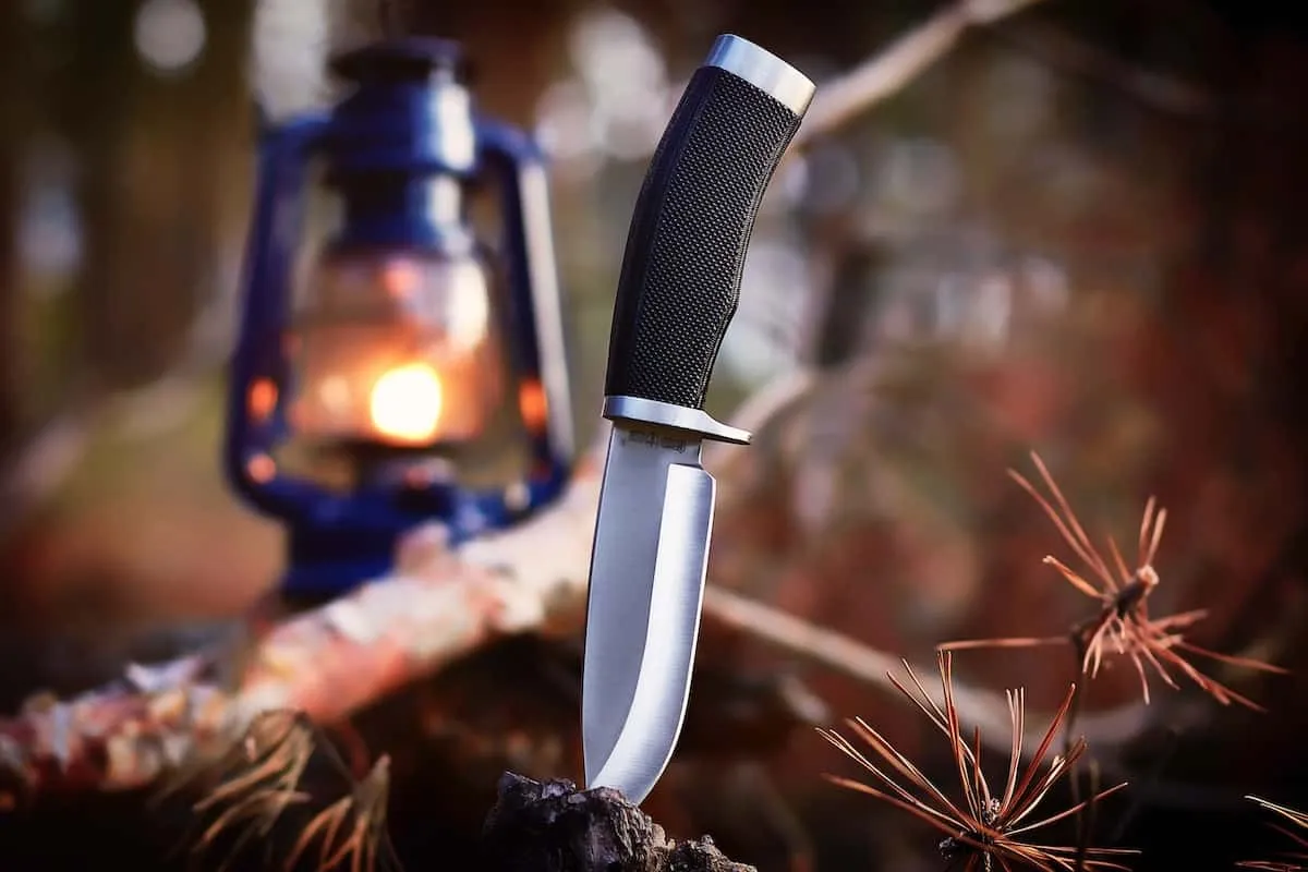 Couteau fixe drop-point planté dans une bûche, lanterne à pétrole allumée en forêt
